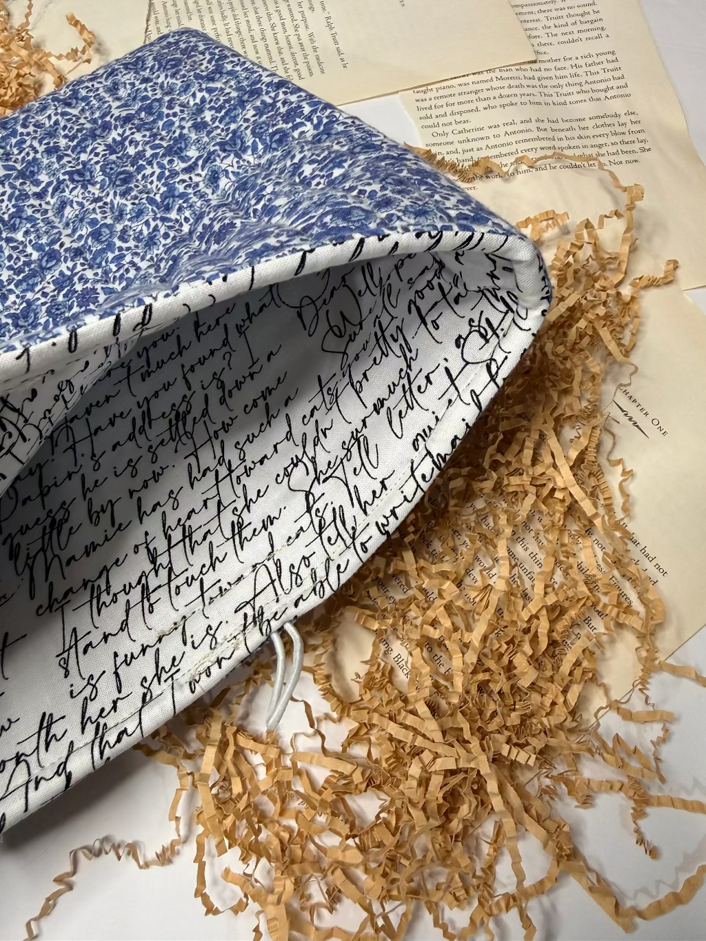 Blue and white floral quilted book sleeve with white interior and black text on  with shredded paper and book pages as decor.